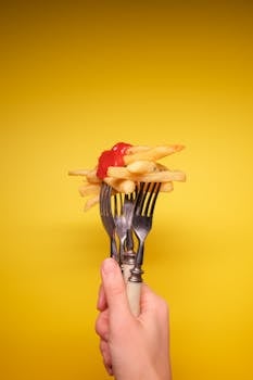 Crop anonymous person hand with forks with fried potatoes and ketchup on yellow background in bright studio