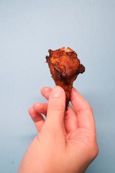 Close-up of a crispy fried chicken drumstick held by a hand against a blue background.