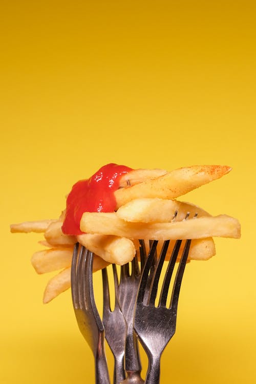 Yummy french fries with ketchup on forks near yellow wall in light studio