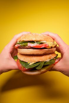 Crop anonymous person hands with burgers with tomatoes and green leaves with meat cutlet on yellow background in bright studio