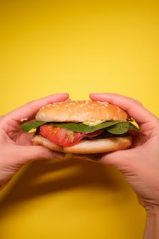 Crop faceless person hands with burger with tomatoes and green leaves with meat cutlet on yellow background in bright studio
