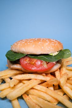 Burger with tomatoes slices and green leaves with meat cutlet on french fries on blue surface in light studio