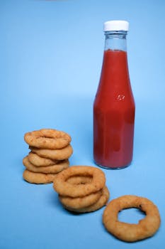 Tasty golden deep fried onion rings near bottle with ketchup in light studio on blue surface