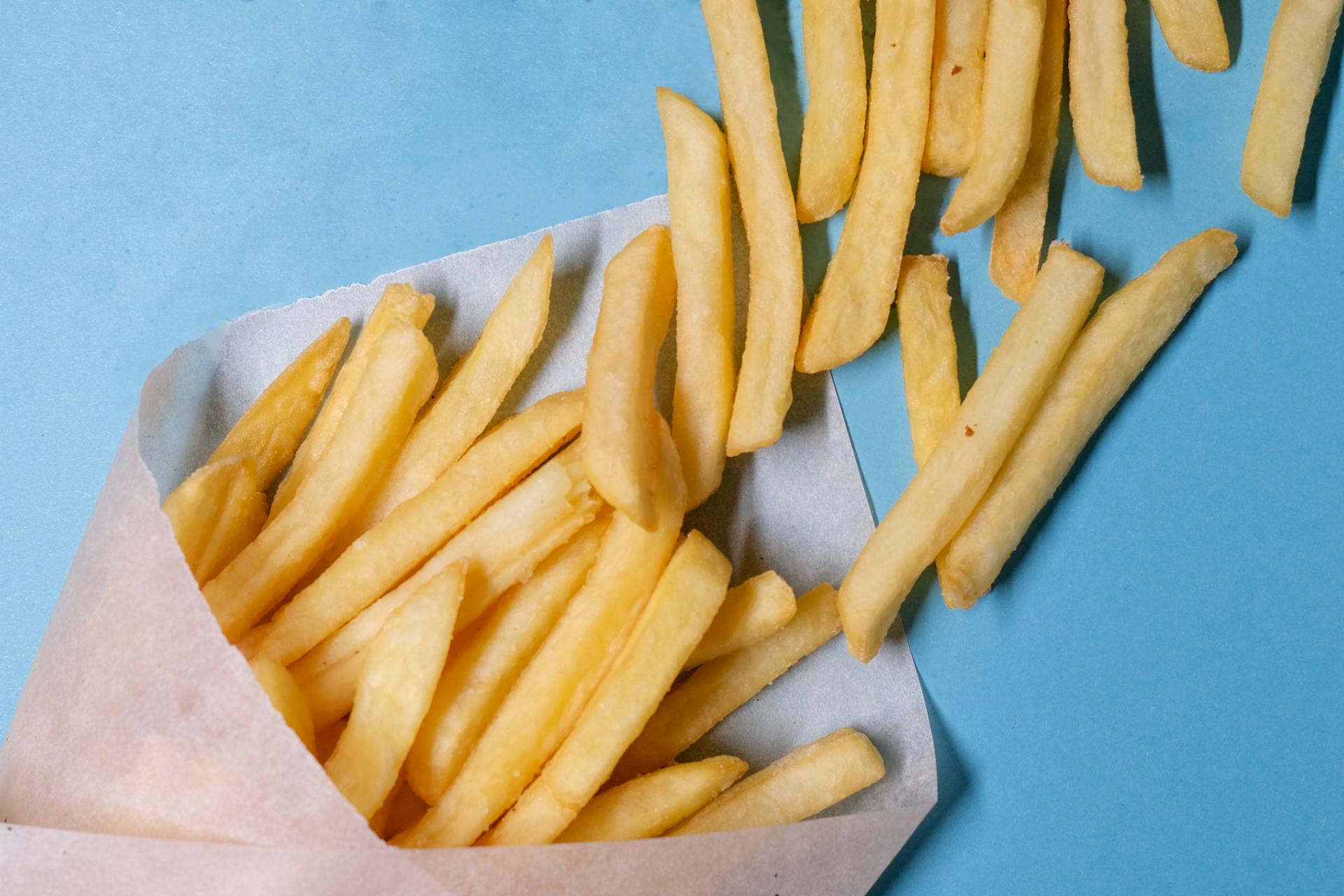 Top view of tasty french fries in paper placed on blue table in light studio