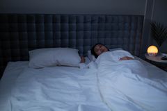 An Asian woman lying awake under soft white bedding in a dimly lit modern bedroom.
