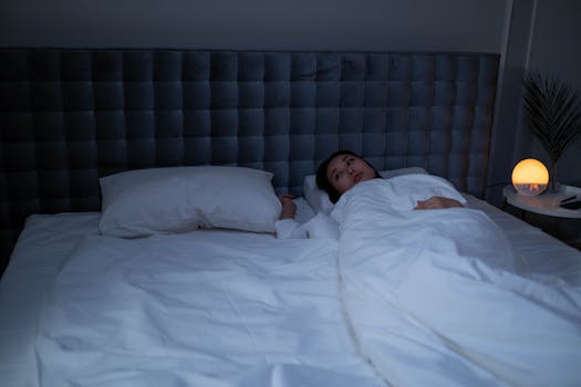 An Asian woman lying awake under soft white bedding in a dimly lit modern bedroom.
