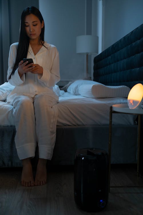 Free Woman in White Pajamas Sitting on Bed While Using a Phone Stock Photo