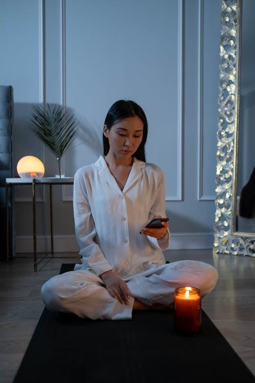 Woman in White Pajamas Sitting on a Yoga Mat Using Her Cell Phone