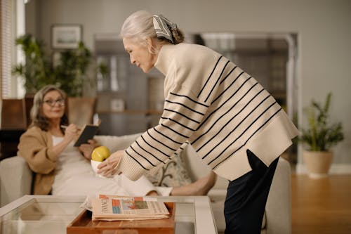 Free An Elderly Woman Sitting on a Couch while Looking at the Elderly Woman Standing Beside the Table while Holding a Bowl of Fruit Stock Photo