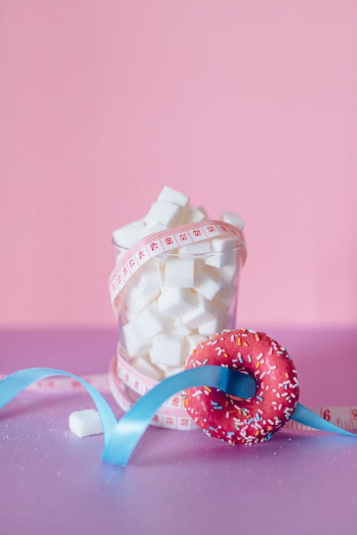 Blocks of Sugar on a Glass Beside a Donut with Blue Ribbon on Pink Surface