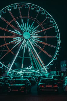 A glowing Ferris wheel towering above parked cars at night in a cityscape, creating a vibrant urban ambiance.