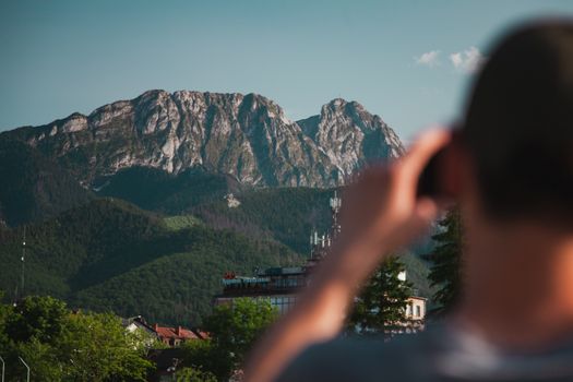 Back view of anonymous male taking photo of buildings near green trees and plants with stone hills on background under blue sky in daytime in countryside