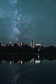Still water against starry sky and multistory glowing residential houses located on shore near lush trees in city at night time