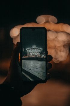 Unrecognizable person taking picture of merry go round with glowing lights on smartphone on blurred background at night time in amusement park