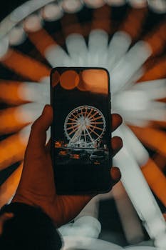 Crop unrecognizable male traveler using smartphone while taking photo of illuminated Ferris wheel in amusement park at night
