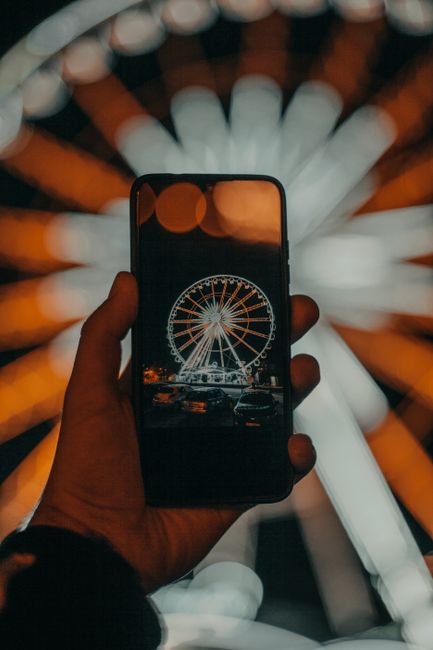 Crop unrecognizable male traveler using smartphone while taking photo of illuminated Ferris wheel in amusement park at night