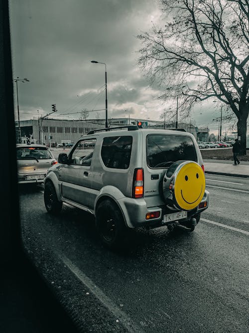 Through window of modern SUV car with smiley face sticker on spare wheel driving on asphalt road against cloudy sky in city