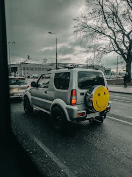 Through window of modern SUV car with smiley face sticker on spare wheel driving on asphalt road against cloudy sky in city