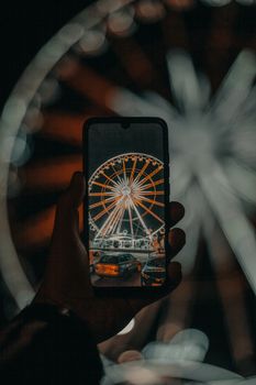 Silhouette of crop anonymous male tourist taking photo of illuminated Ferris wheel on mobile phone in amusement park at night