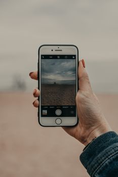 Crop unrecognizable female traveler holding smartphone while taking photo of picturesque sea under cloudy sky on sandy beach