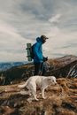 Side view of unrecognizable young male backpacker in warm clothes ascending on rough rocky mountain with trekking poles during hiking trip with adorable Labrador Retriever dog