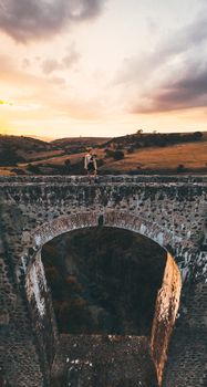 A lone traveler stands atop an ancient stone bridge at sunset, showcasing breathtaking scenery.