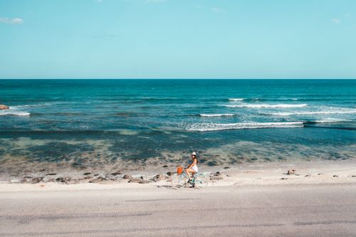 Free A woman rides a bicycle along a scenic beachside road under a clear blue sky. Stock Photo