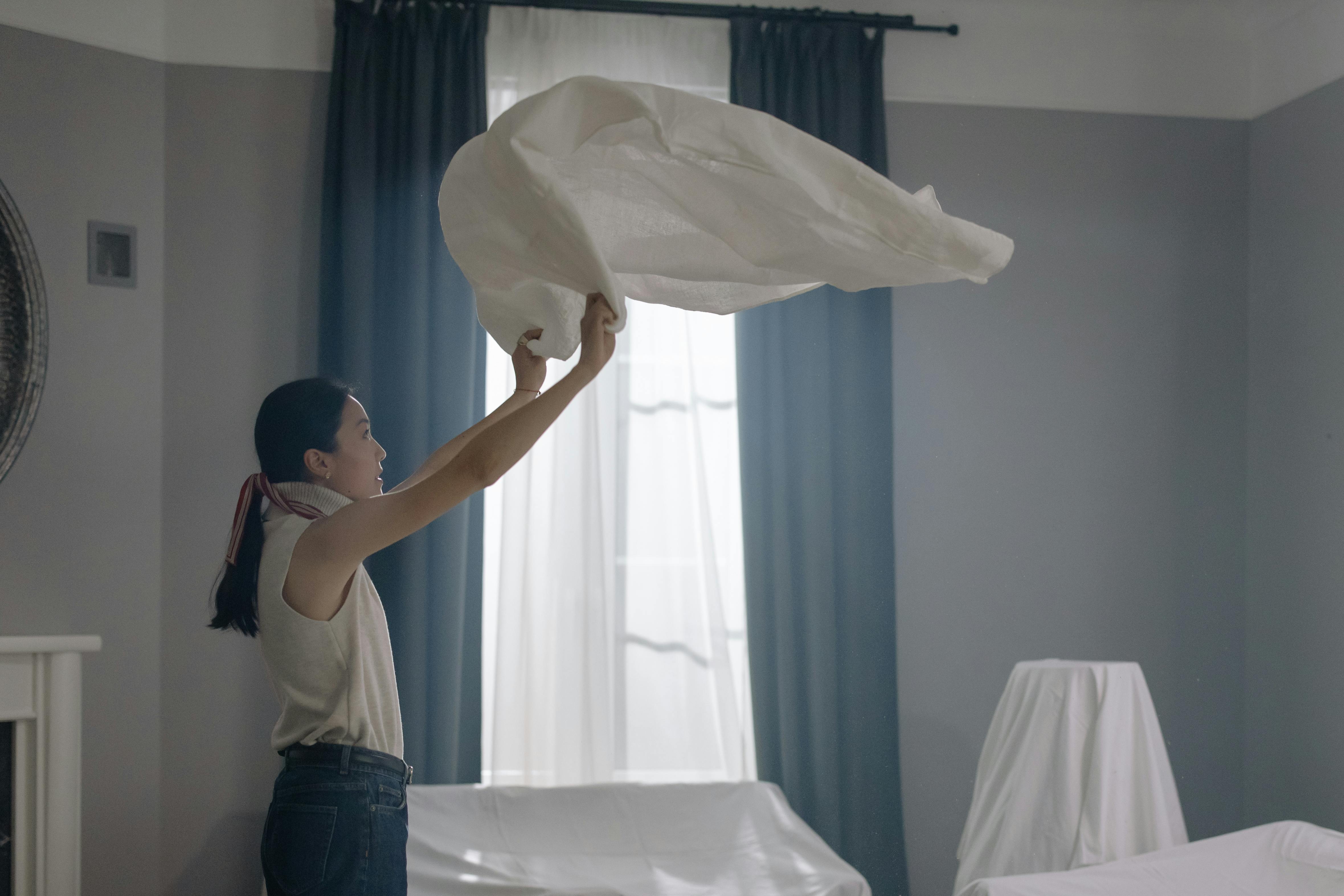An Asian woman in a sleeveless shirt shakes out a white sheet in a well-lit room.