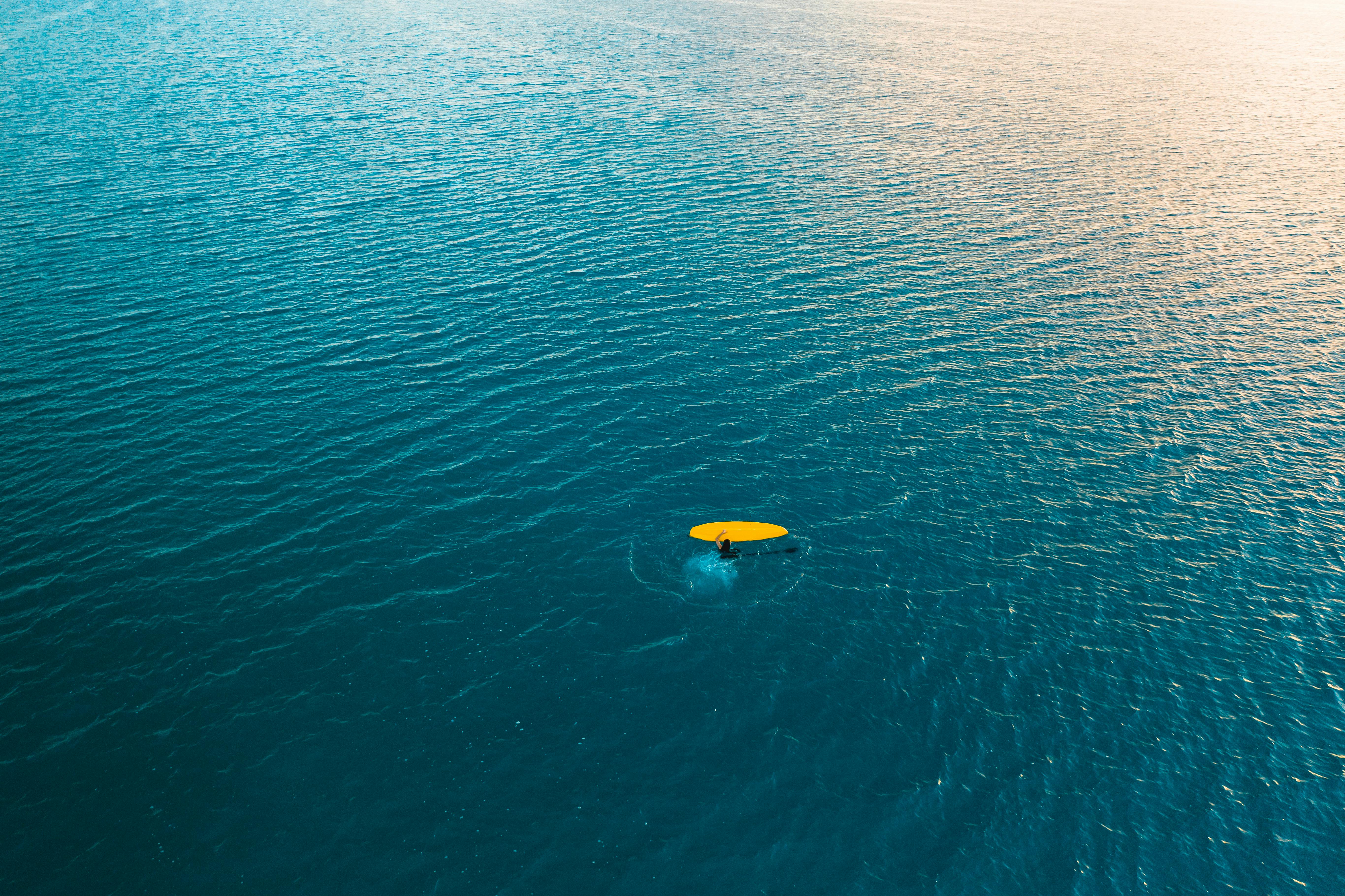 Drone captures surfer paddling on a vast, calm blue ocean under clear daylight skies.