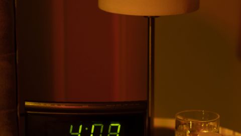 A warm and moody bedside table with a digital clock showing 4:08 and a lamp in dim lighting.