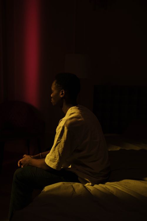 Free A contemplative man sits alone in a shadowy bedroom with a warm dim light. Stock Photo