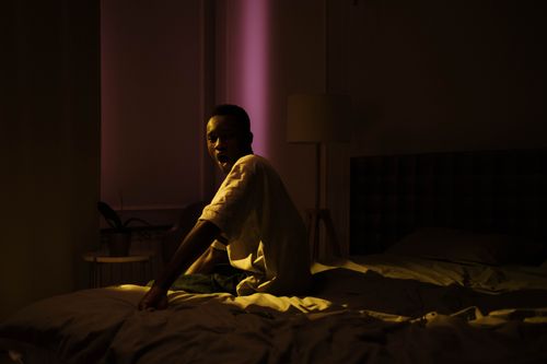 Free A man sits on a bed in a dimly lit bedroom, with moody lighting and deep shadows. Stock Photo