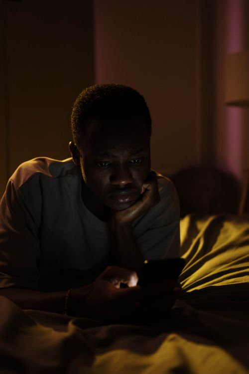 Free A man lying in bed using his smartphone in a dimly lit room, conveying a nighttime mood. Stock Photo