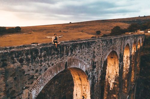 Free Drone view of a woman sitting on a historic aqueduct in a rural landscape during a cloudy day. Stock Photo