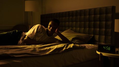 A man lies awake in bed, illuminated by early morning light from a clock reading 4:56 AM.