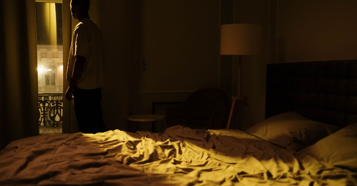 Man in a dark bedroom gazing out the window at night
