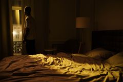 Man in dark bedroom gazing out window, highlighting themes of insomnia and solitude.
