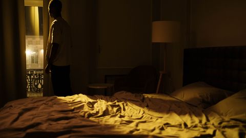 Man in dark bedroom gazing out window, highlighting themes of insomnia and solitude.
