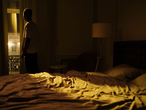 Man in dark bedroom gazing out window, highlighting themes of insomnia and solitude.