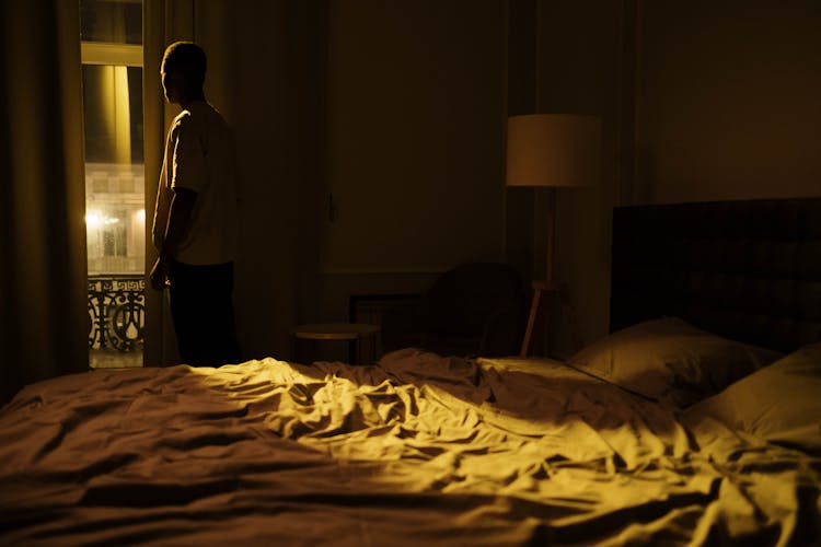 Young Man Standing By Window In Bedroom At Night