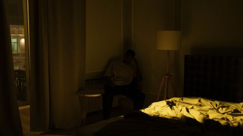 An adult man experiencing insomnia, sitting in a dimly lit bedroom corner at night.