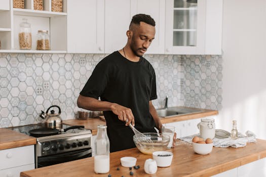 Man preparing pancakes in a contemporary kitchen setting with fresh ingredients on a wooden countertop.