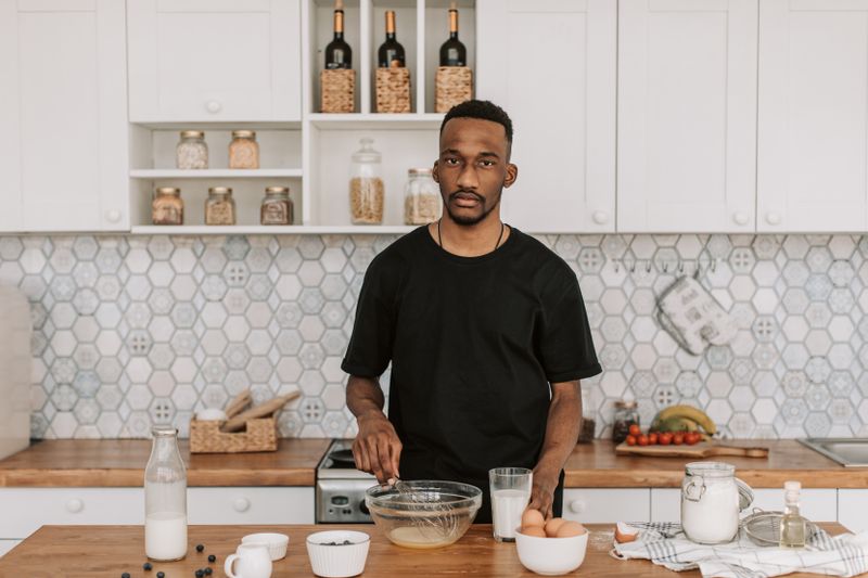 A Black man in a black T-shirt stands at a kitchen island with a mixing bowl, a bottle of milk, eggs, and a small bowl of berries in front of him.