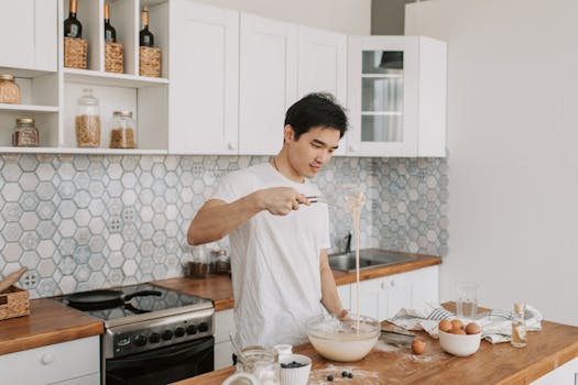 Asian man making batter at home using whisk with eggs and blueberries nearby.