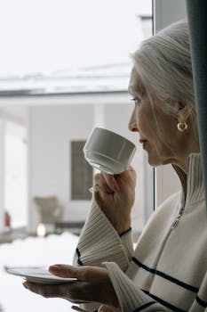 Senior woman with gray hair enjoys a warm coffee, gazing out a snowy window.