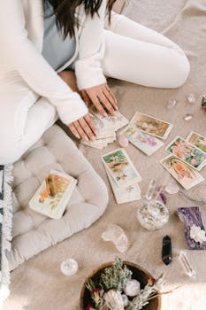 A woman sits cross-legged indoors, handling tarot cards, surrounded by crystals and herbs.