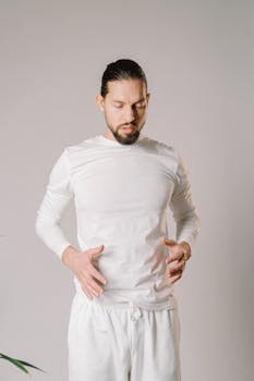 A man with facial hair stands in a contemplative pose, wearing a white outfit.