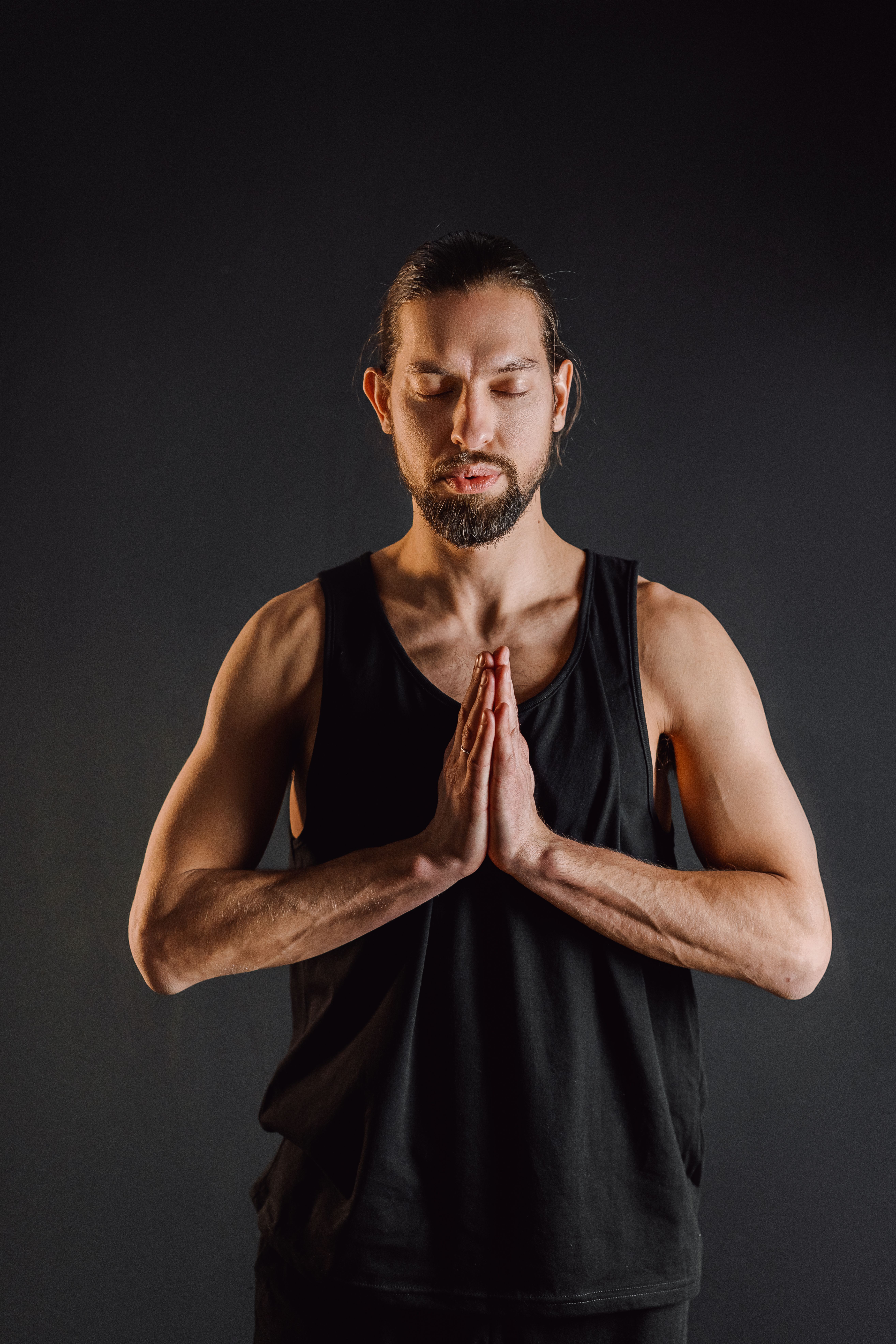 A man in a black tank top meditates with praying hands against a dark background.