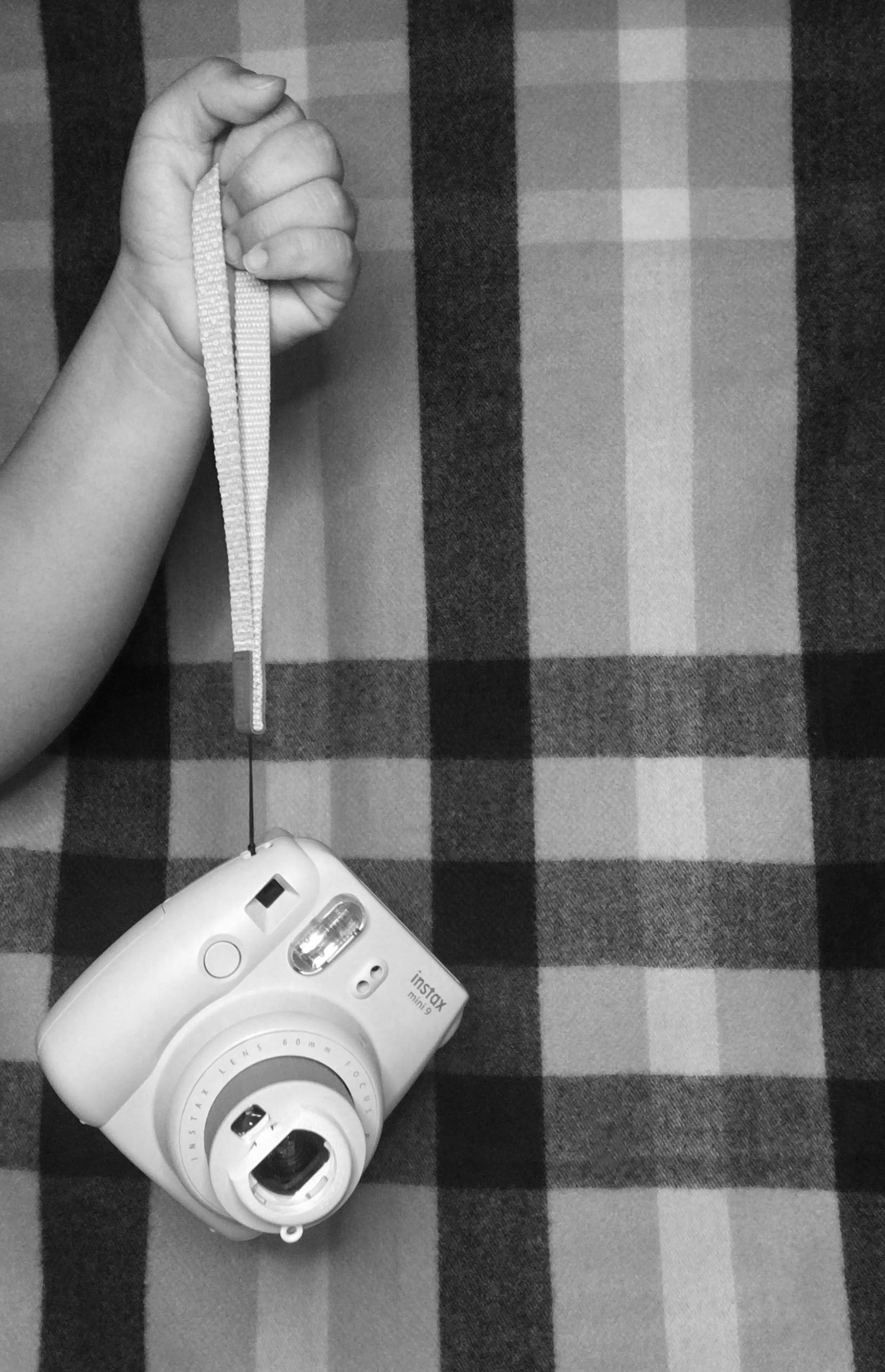 Black and white photo of a hand holding an instant camera against a plaid background.