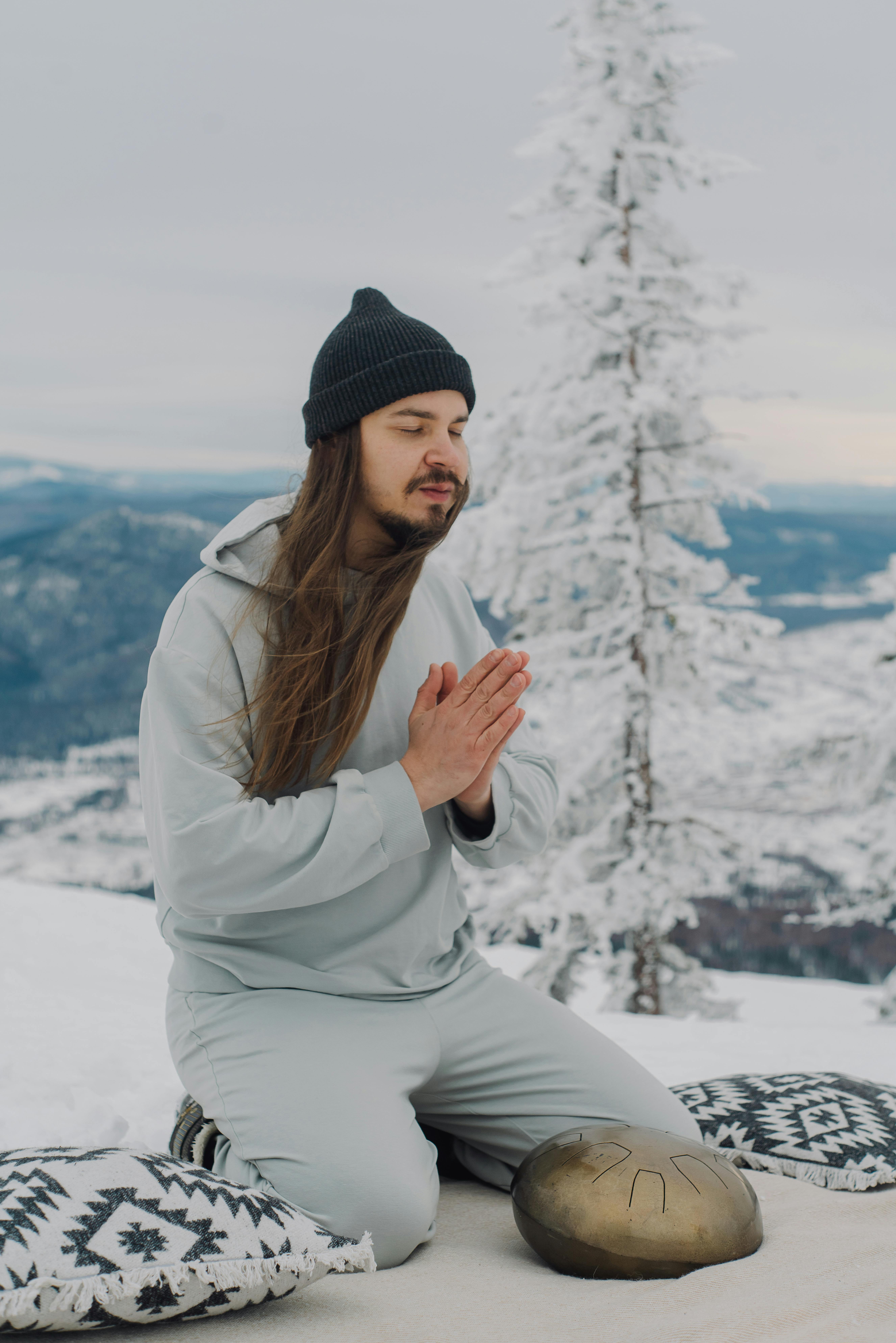 Full body of peaceful male with namaste gesture sitting on knees on snowy ground near steel tongue drum during meditation in highland